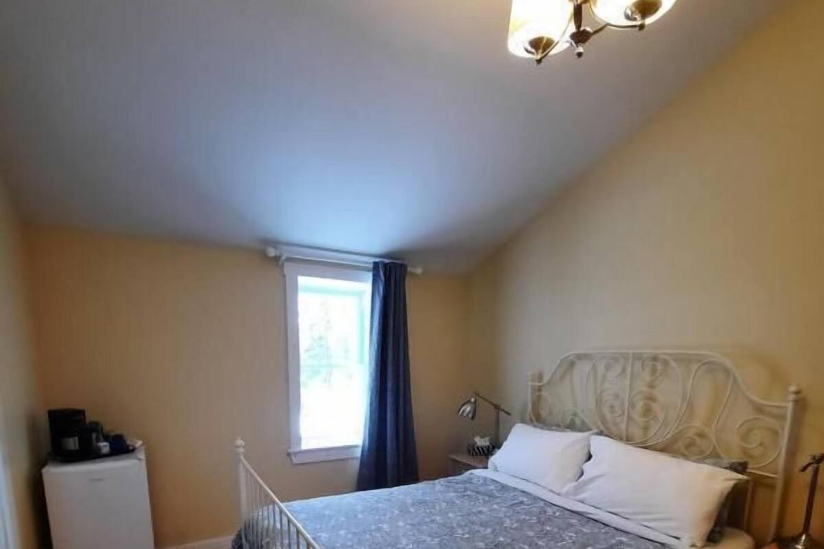A bedroom with a bed, a nightstand, a lamp, a window, and a small refrigerator.