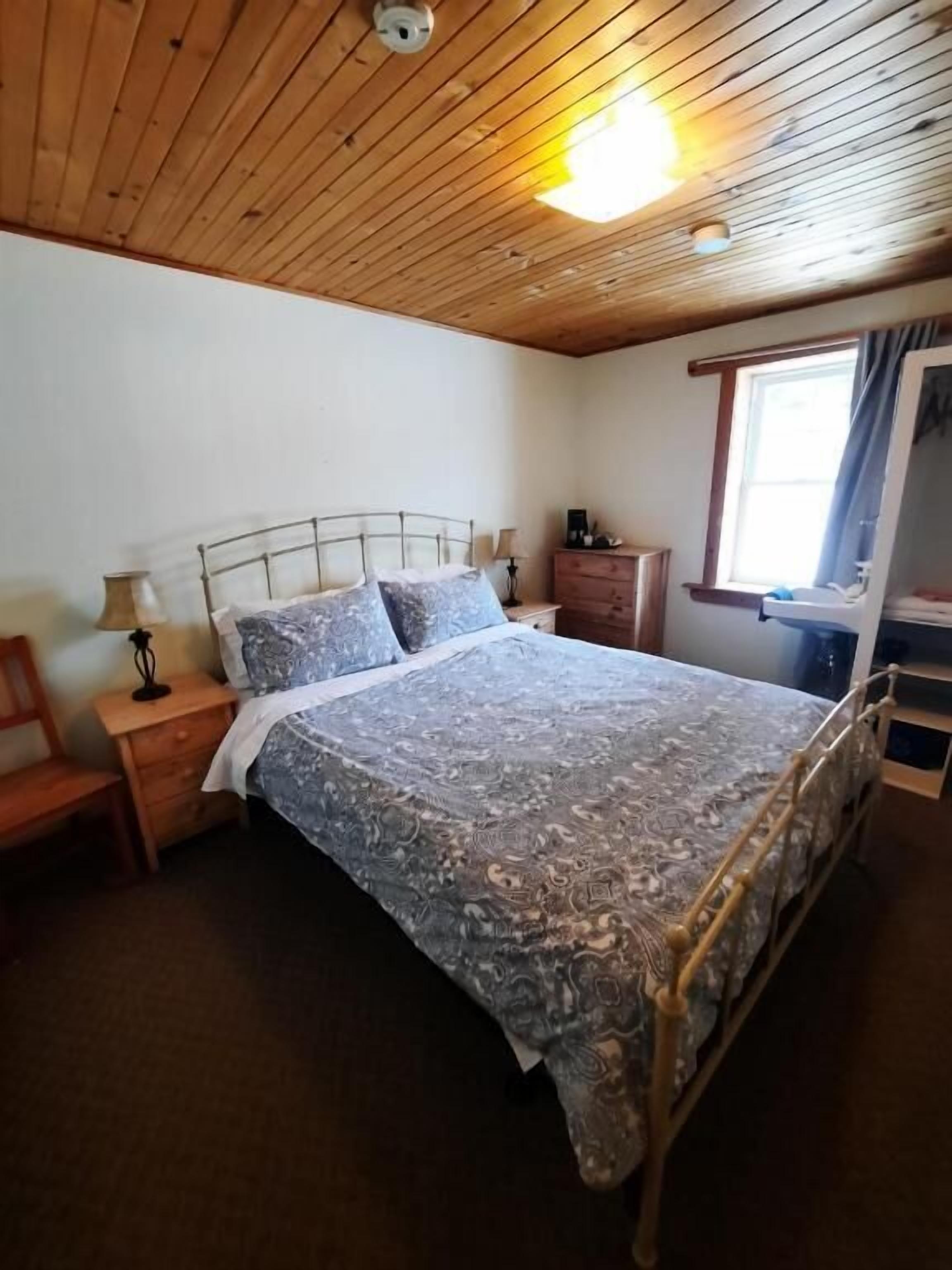 A bedroom with a wooden ceiling, a metal bed frame, a patterned bedspread, a nightstand, a dresser, and a window with curtains.