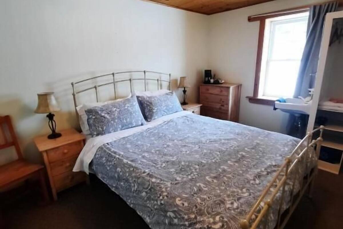 A bedroom with a wooden ceiling, a metal bed frame, a patterned bedspread, a nightstand, a dresser, and a window with curtains.