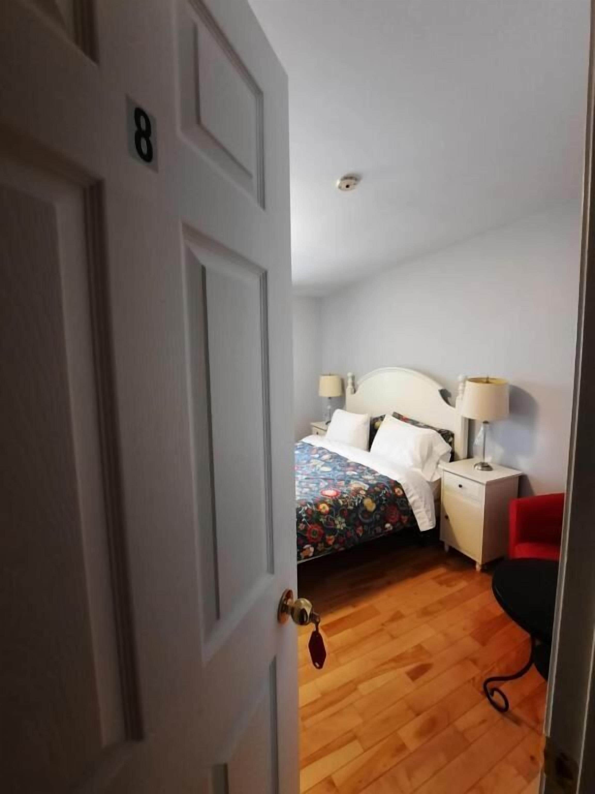 Una habitación con una cama, lámparas de noche, una silla roja y suelo de madera.
