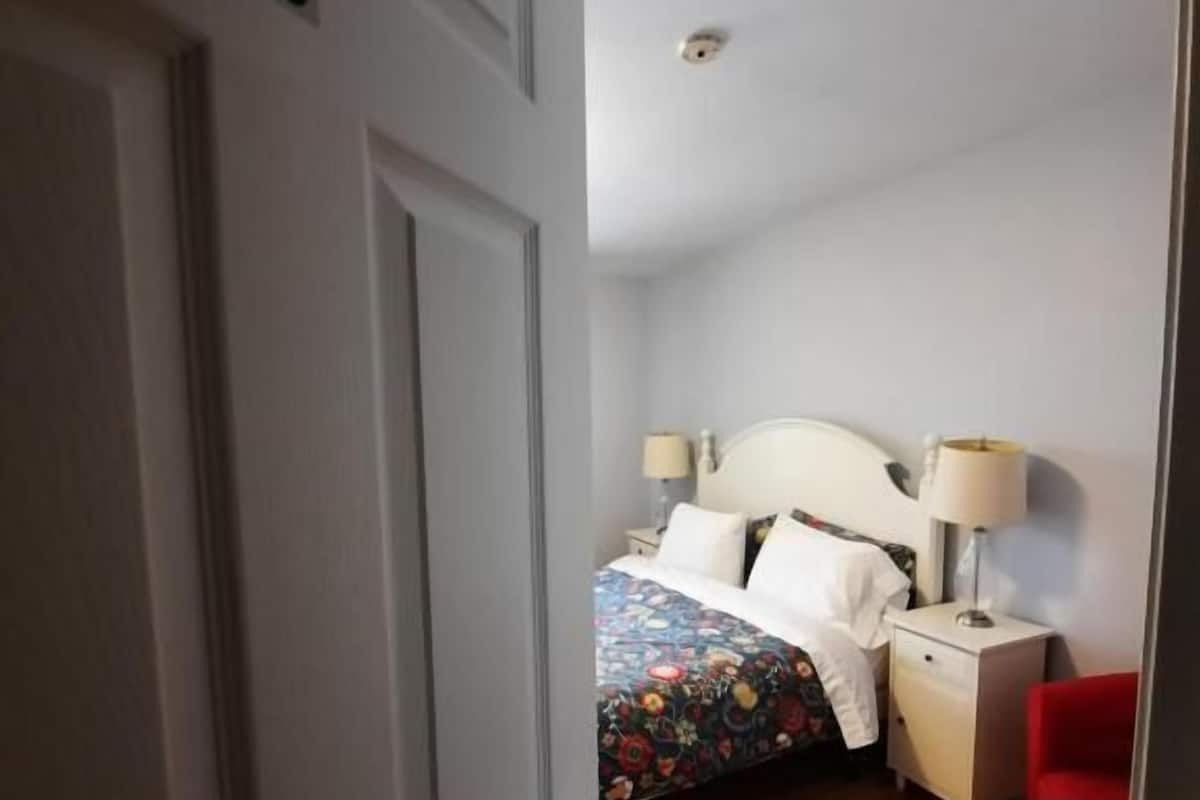 Una habitación con una cama, lámparas de noche, una silla roja y suelo de madera.