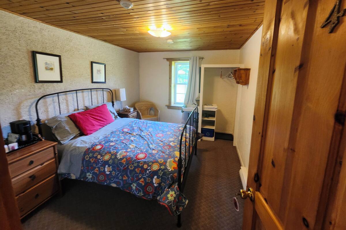 A bedroom with a wooden ceiling, a bed with a floral bedspread, a nightstand, a chair, a window with curtains, and a wooden wardrobe.