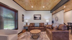 A living room with a wooden ceiling, a brown leather sofa, a round coffee table, and a large window with blinds. - *Charming 4BD Lodge Near TOWN! Fire Pit + Hot Tub* (Sevierville)