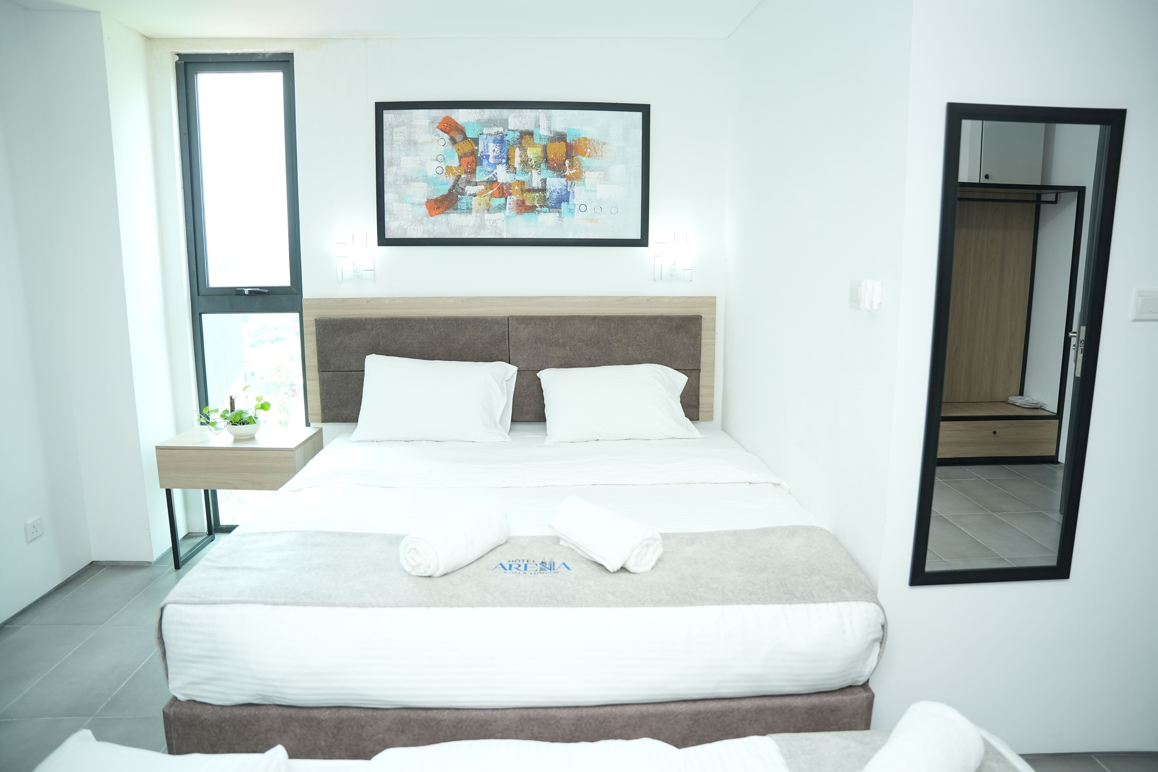 A modern hotel room with a large bed, a bedside table with a plant, a framed abstract painting, and a mirror on the wall.