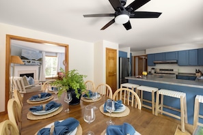 A dining area with a wooden table set for four, a ceiling fan, and a kitchen with blue cabinets and a stainless steel refrigerator. - Acadia Escape: Hot Tub, Sauna, Game Room, Fire Pit (Mount Desert)
