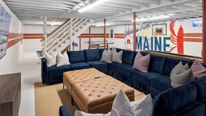 A modern interior with a sectional sofa, a tufted ottoman, and a staircase. - Acadia Escape: Hot Tub, Sauna, Game Room, Fire Pit (Mount Desert)