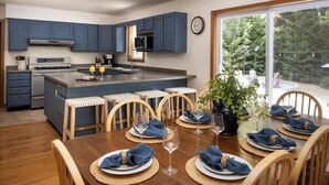 A modern kitchen with blue cabinets, a central island with bar stools, and a dining table set for six with blue napkins and wine glasses. - Acadia Escape: Hot Tub, Sauna, Game Room, Fire Pit (Mount Desert)
