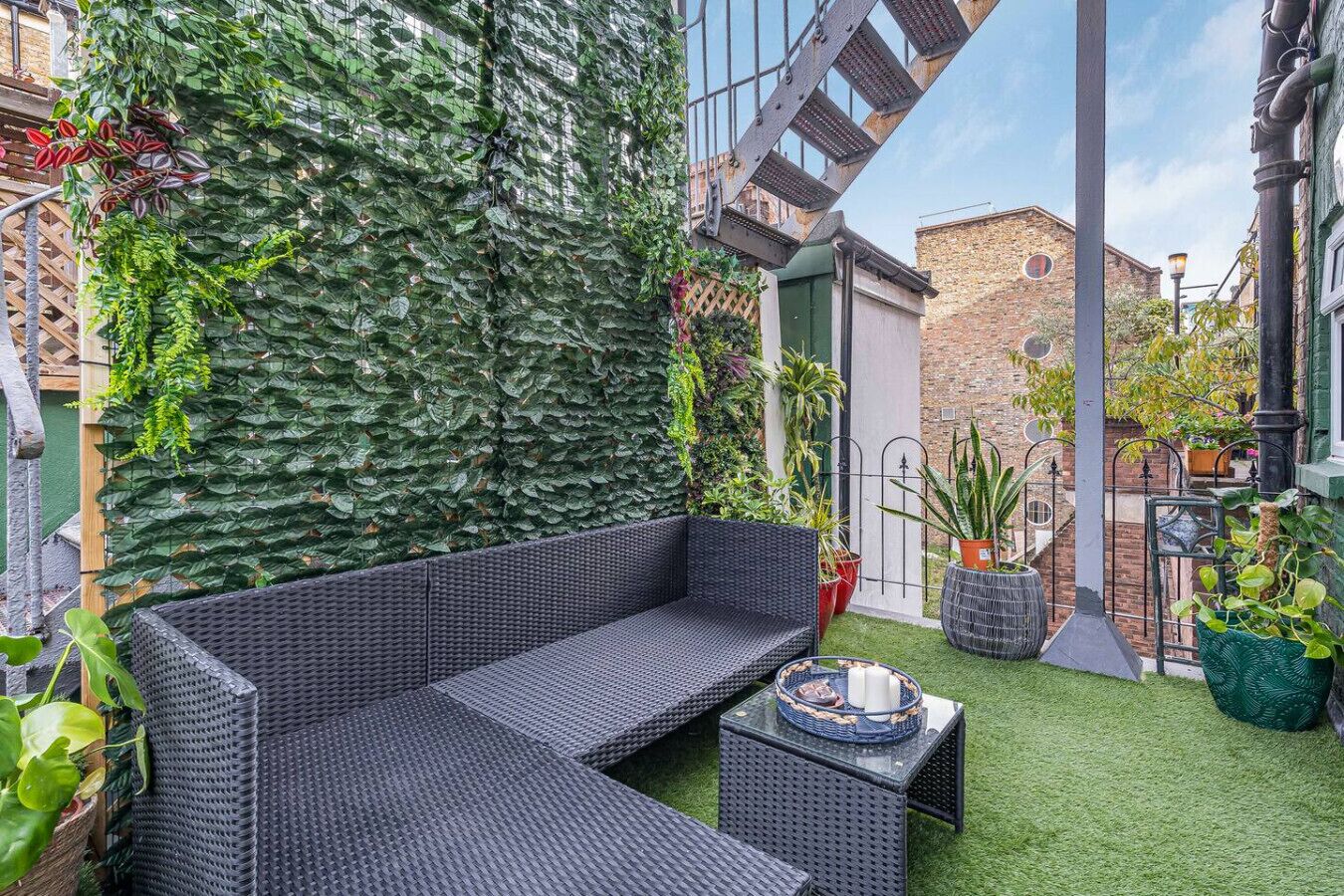 Un espacio al aire libre acogedor con un sofá de mimbre y una mesita pequeña, rodeado de abundante follaje y plantas en macetas.