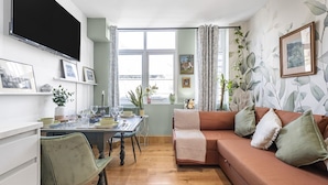 A modern living room with a dining area, featuring a sofa, chairs, a table set for dining, and a large window providing natural light.