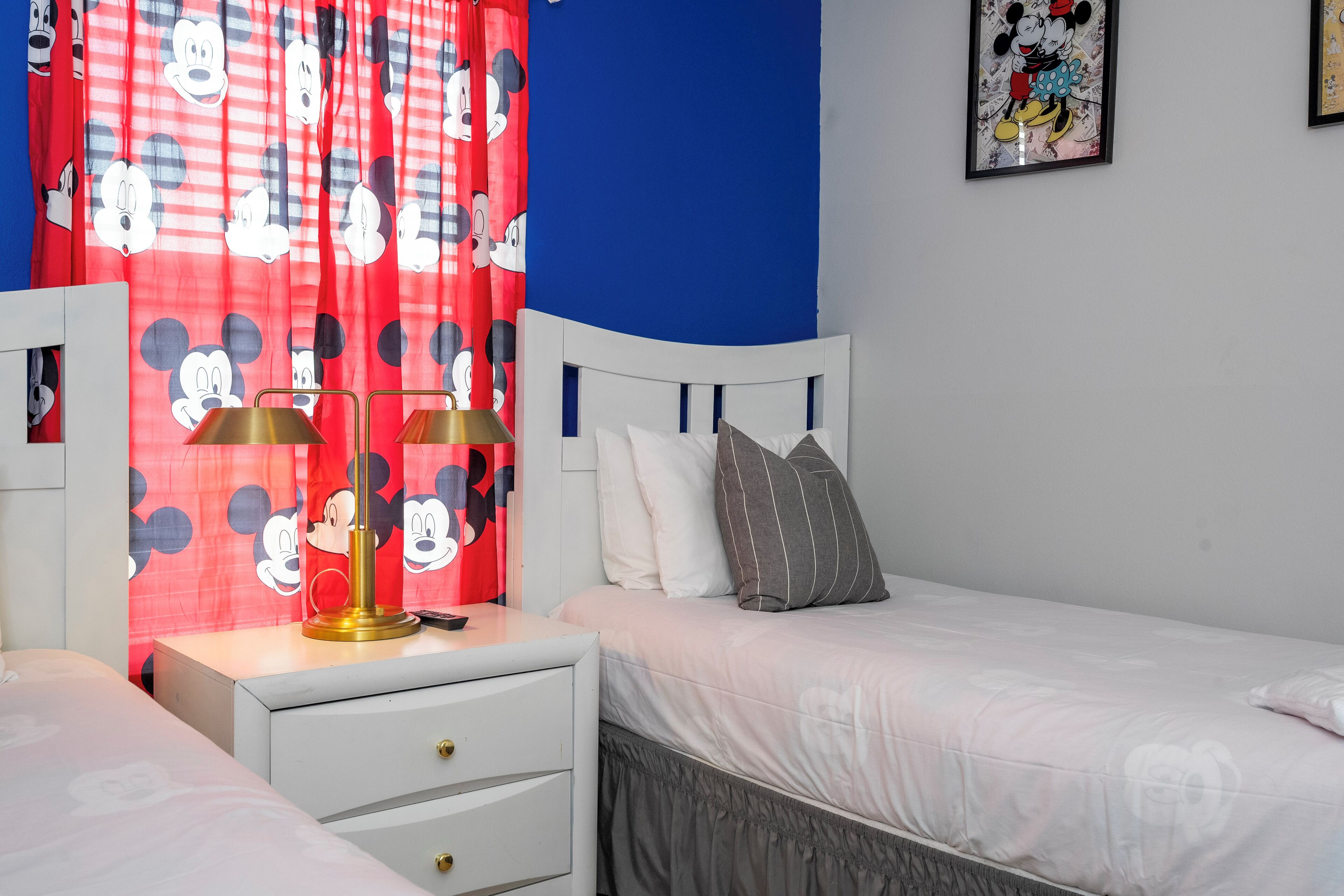 A bedroom with a bed, a nightstand, a lamp, Mickey Mouse curtains, and a wall with a framed picture.