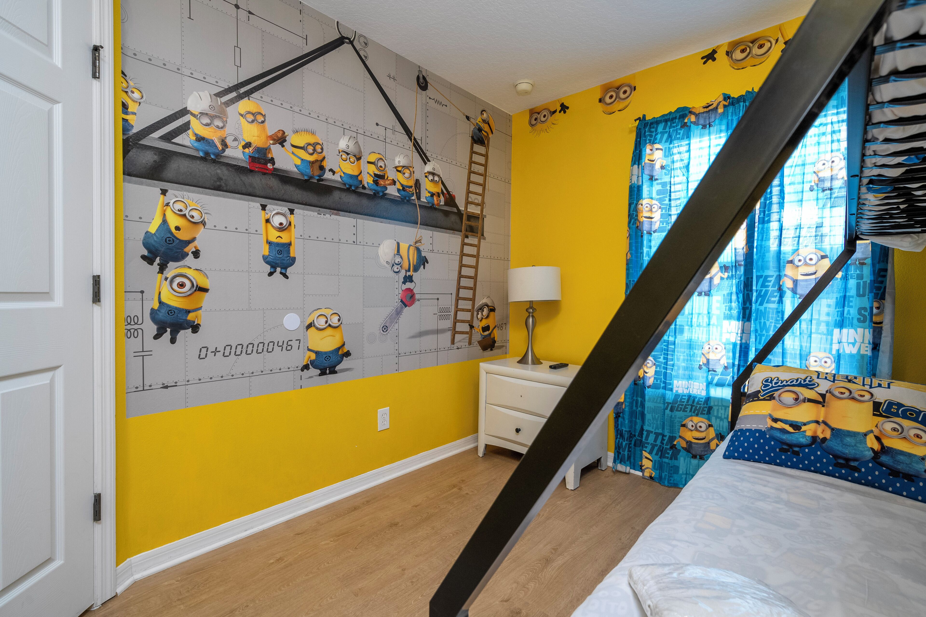 Una cameretta con un murales a tema Minions, un letto, un comodino e una finestra con tende.