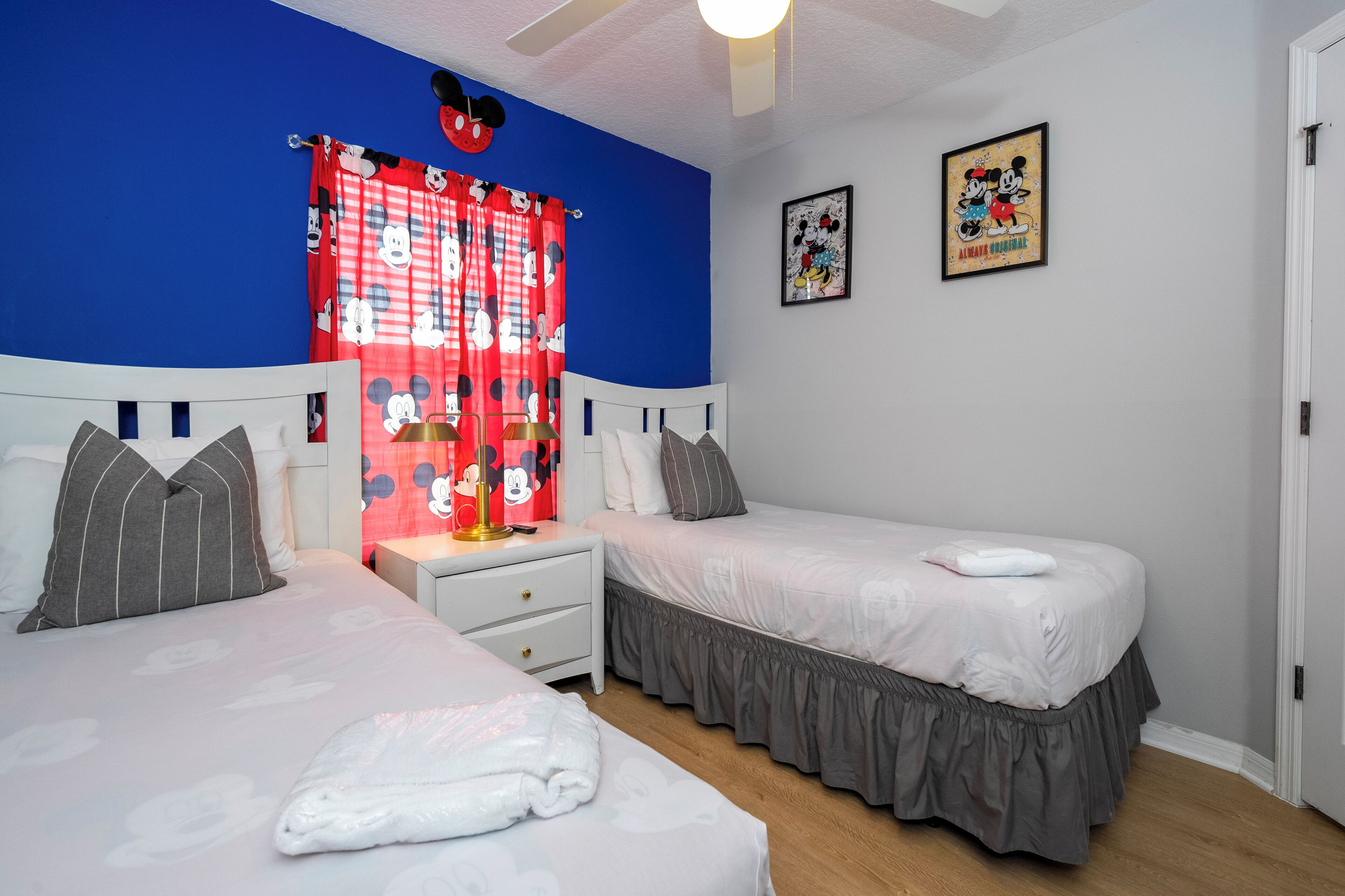 Une chambre avec deux lits, une table de chevet, un rideau à l’effigie de Mickey Mouse et deux tableaux encadrés accrochés au mur.