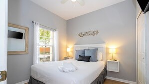 A neatly made bed with a white and gray bed skirt, two bedside lamps, a wall-mounted TV, and a window with blinds. - 4BR Bella Vida Villa with Pool, BBQ & Themed Rooms near Disney (Kissimmee)