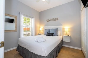 Un lit bien fait, avec un couvre-lit blanc et gris, deux lampes de chevet, un téléviseur fixé au mur et une fenêtre avec des stores.
