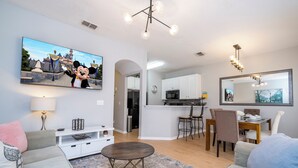 A modern living room with a flat-screen TV, a dining area, kitchen, and a large mirror. - 4BR Bella Vida Villa with Pool, BBQ & Themed Rooms near Disney (Kissimmee)