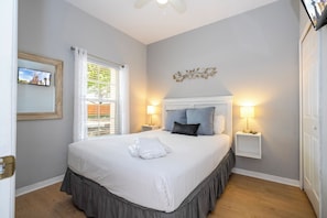 A neatly made bed with a white comforter and a skirted bed skirt, two bedside lamps, a framed picture on the wall, and a window with blinds.