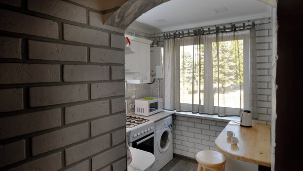 A compact kitchen with a brick wall, a window with curtains, a microwave, a stove, and a washing machine. - KAYARAN home (Gyumri)