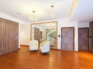 A modern hotel lobby with wooden flooring, two white armchairs, a glass partition with a geometric design, and two wooden doors.