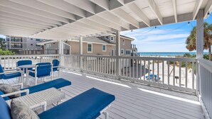 A covered outdoor deck with blue furniture, a white railing, and a view of the beach.