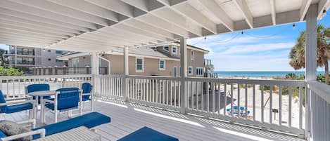 A covered outdoor deck with blue furniture, a white railing, and a view of the beach.