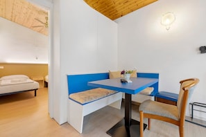 Une pièce comprenant un lit, un banc bleu, une chaise en bois, une table avec une télécommande et un luminaire fixé au mur.