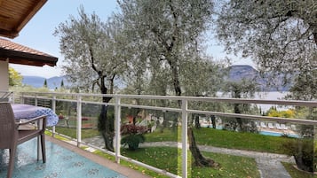 Un balcone con tavolo e sedie, vista su una piscina e un giardino alberato con montagne sullo sfondo.