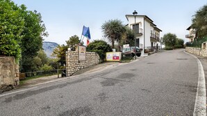A road leading to a building with a stone wall, a flag, and a signboard. - Apartment 'Bungalow Pit Stop' with Lake View, Wi-Fi and Air Conditioning (Brenzone sul Garda)