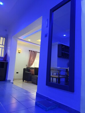 Una sala de estar con un sofá, una mesa de comedor con sillas, un espejo grande y una escalera que conduce a un piso superior.
