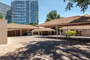 Un espace de stationnement doté d’abris pour voitures, d’un chemin piétonnier pavé et, au fond, d’un bâtiment avec un toit recouvert de tuiles.