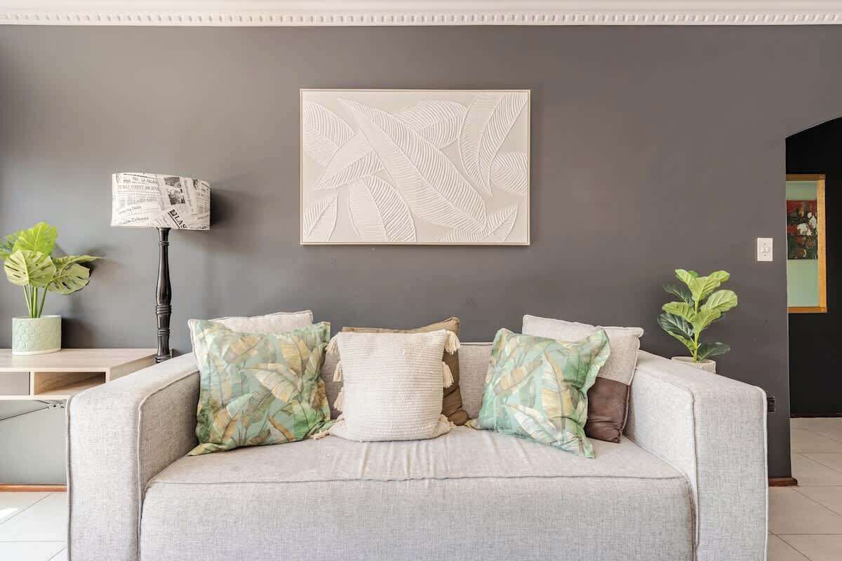Un salón con un sofá gris, cojines estampados, una obra de arte colgada en la pared y dos plantas en macetas.