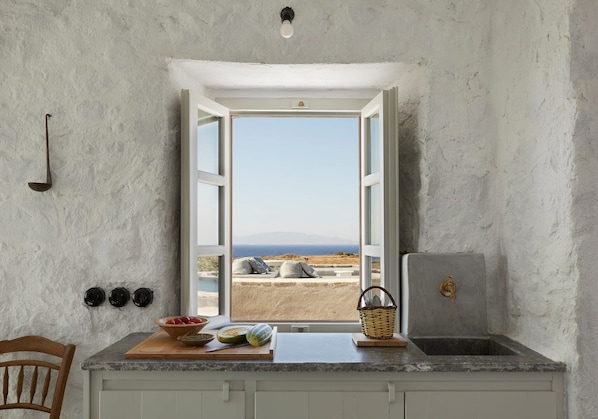 A kitchen with a view of the sea, a wooden chair, a bowl of fruit, and a basket on the counter.