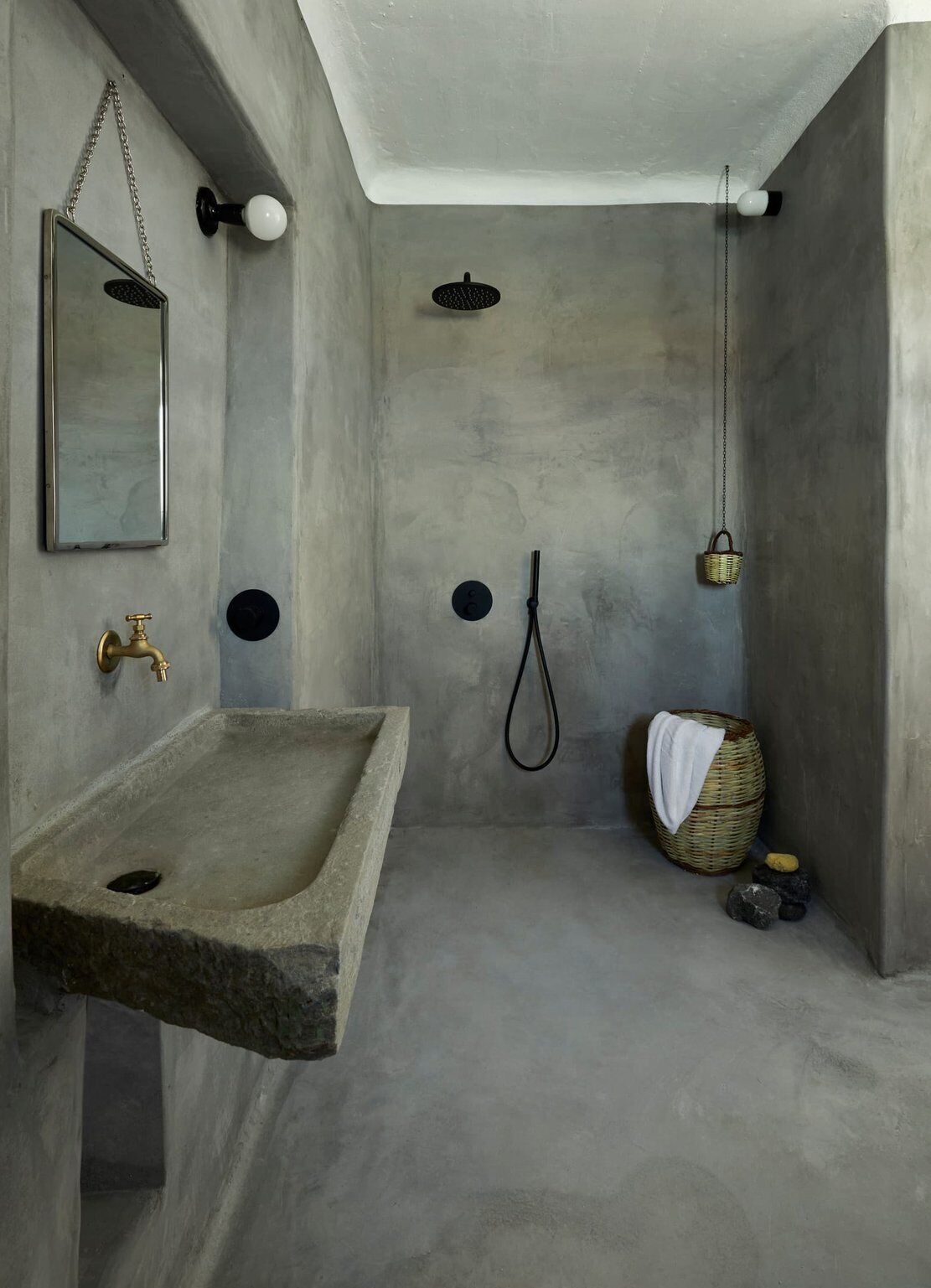 A minimalist bathroom with a stone sink, a mirror, a shower with a handheld showerhead, and a small basket with a towel.