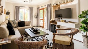 A modern living room with a sofa, coffee table, wicker chairs, a flat-screen TV, and a large window with curtains. - Balcony in Trees|1km to  Mandela Sq (Sandton)