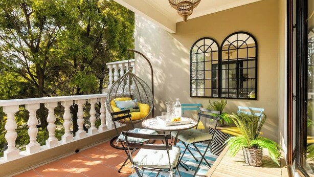 A balcony with a white balustrade, a round table with chairs, a hanging chair, and a window with black frames. - Balcony in Trees|1km to  Mandela Sq (Sandton)