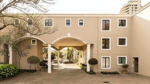 A building with a central archway, multiple windows, and a paved courtyard with greenery. - Balcony in Trees|1km to  Mandela Sq (Sandton)