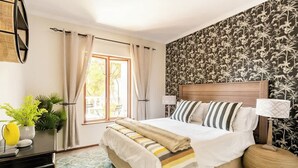 A bedroom with a bed, curtains, a window, a desk, and a wall with a floral pattern. - Balcony in Trees|1km to  Mandela Sq (Sandton)
