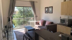 A compact living space with a kitchenette, a sofa, a dining table, and a balcony with a view of greenery.