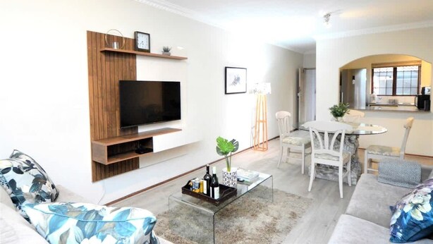 Una sala de estar moderna con televisor de pantalla plana, estanterías de madera, una mesa de centro con bebidas, un sofá y un comedor con sillas.