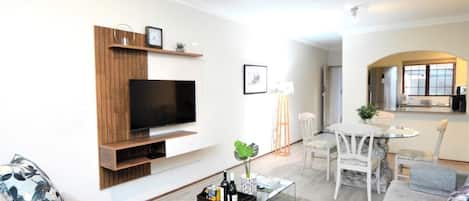 A modern living room with a flat-screen TV, wooden shelving, a coffee table with drinks, a sofa, and a dining area with chairs.