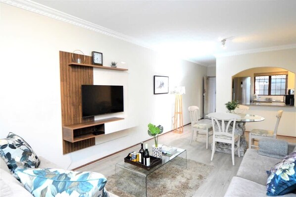 A modern living room with a flat-screen TV, wooden shelving, a coffee table with drinks, a sofa, and a dining area with chairs. - Quiet Garden Apartment | Heart of Sandton (Sandton)