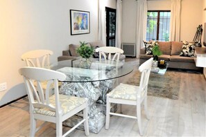 A modern living room with a glass-top dining table surrounded by white chairs, a sofa, and a coffee table with decorative items.