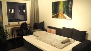 A hotel room with a bed, a desk, a chair, and a painting of a forest path. - Central apartment with secure private parking in the backyard (Ludwigshafen am Rhein)