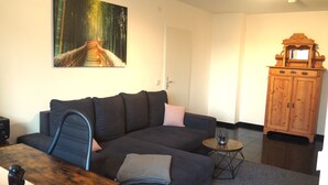 A living room with a dark-colored sofa, a small round table with a lamp, a wooden cabinet, and a painting of a forest scene on the wall. - Central apartment with secure private parking in the backyard (Ludwigshafen am Rhein)