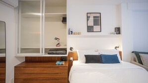 A modern hotel room with a wooden nightstand, a bedside table, a bed with white bedding, a large window with blinds, a framed artwork on the wall, and a small shelf with decorative items. - For You Moema (São Paulo)