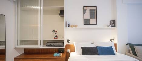 A modern hotel room with a wooden nightstand, a bedside table, a bed with white bedding, a large window with blinds, a framed artwork on the wall, and a small shelf with decorative items.