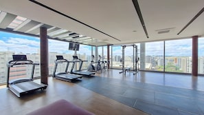 A modern gym with treadmills, elliptical machines, and weightlifting equipment, large windows offering a city view. - For You Moema (São Paulo)