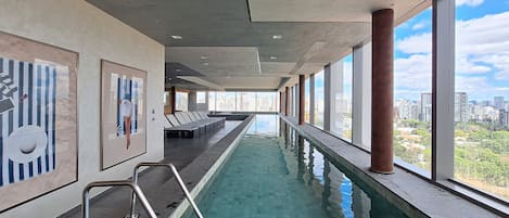 A long indoor swimming pool with a city view, sun loungers, and a modern architectural design.