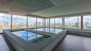 A modern indoor pool with a city view, large windows, and a minimalist design. - For You Moema (São Paulo)