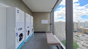 A laundry room with three washing machines, a bench, and a view of the city through large windows. - For You Moema (São Paulo)