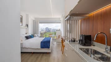 Quarto de hotel moderno com cama, pequena área de refeições, cozinha compacta e grande janela com vista para a cidade.